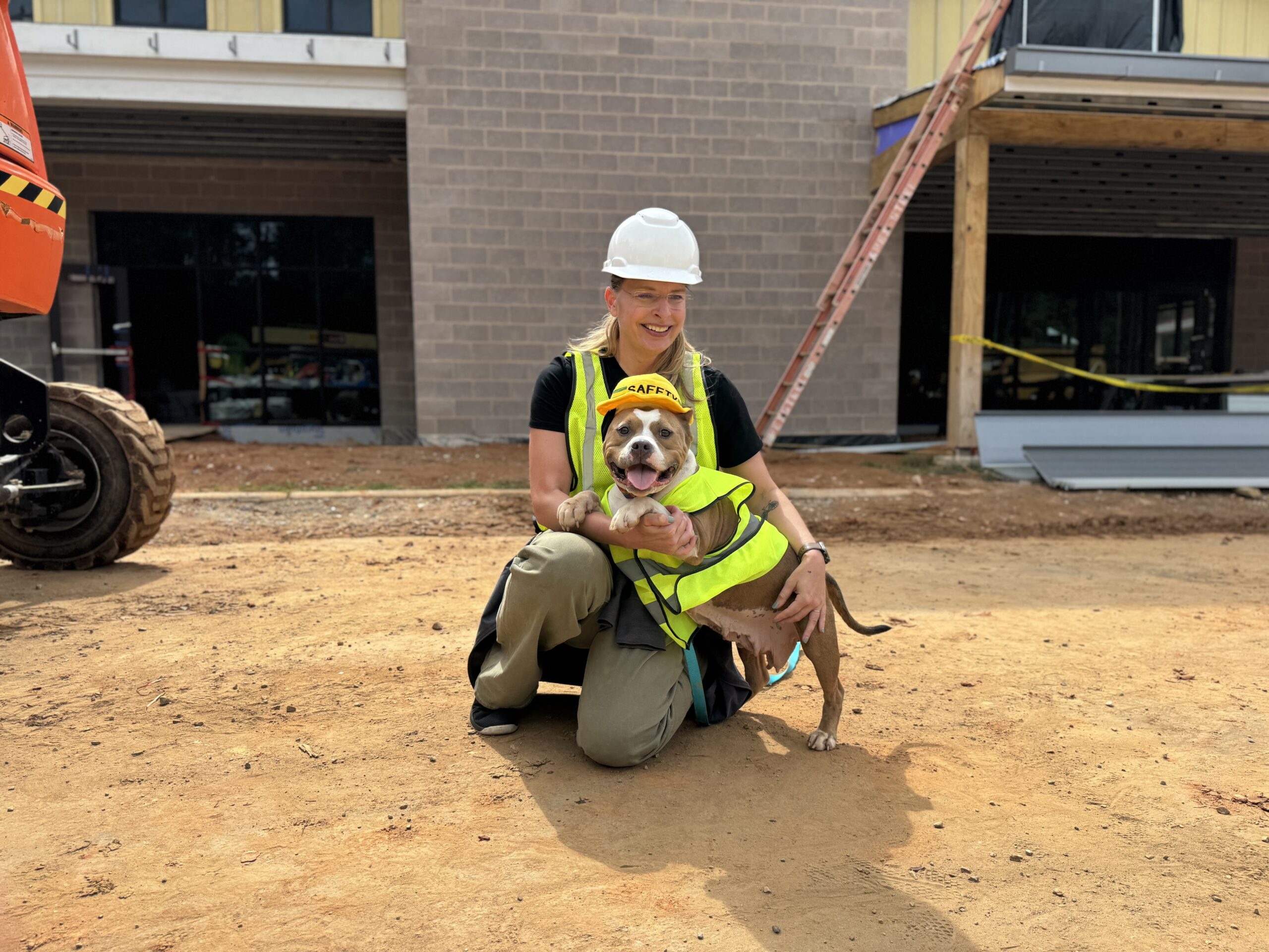 dog and woman both wearing construction vests and hard hats smile and sit outside the construction site of the new animal shelter facility